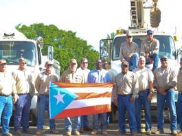Nine electric company employees standing in front of utility trucks holding a puerto rican flag