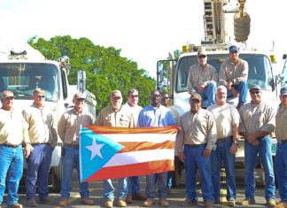 Nine electric company employees standing in front of utility trucks holding a puerto rican flag