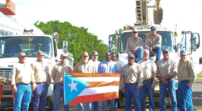 Nine electric company employees standing in front of utility trucks holding a puerto rican flag