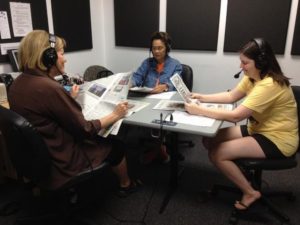 three women sitting in a recording studio wearing headphones and reading newspapers