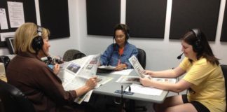 three women sitting in a recording studio wearing headphones and reading newspapers