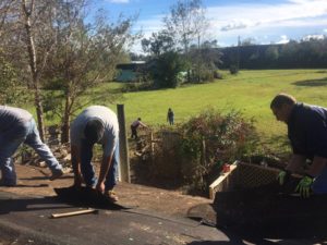 Men tearing shingles off a roof while women cleaning yard in the background