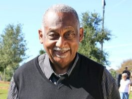 An older man in a black vest standing in a park.