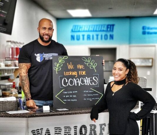 couple standing in front of counter and sign that says "warrior fit, we are looking for need coaches"