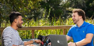 two people sitting at an outdoor table smiling with laptops between them