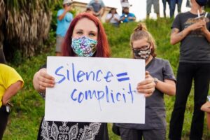 woman holding a sign that says silence - complicit