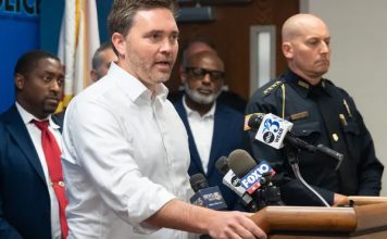 Pensacola Mayor D.C. Reeves speaking at a podium standing next to Pensacola Police Chief Eric Winstrom and city council members