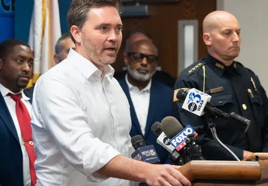 Pensacola Mayor D.C. Reeves speaking at a podium standing next to Pensacola Police Chief Eric Winstrom and city council members
